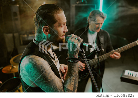 Medium closeup of bearded rock vocalist singing song on stage, mature man playing bass guitar in background Medium closeup of bearded rock vocalist singing song on stage, mature man playing bass guitar in background 116588828