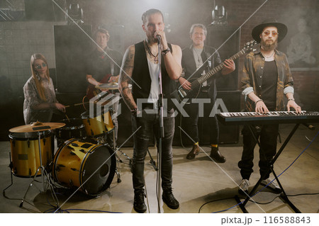 Long shot of modern rock band ready to perform on stage posing for camera with microphone and musical instruments Long shot of modern rock band ready to perform on stage posing for camera with microphone and musical instruments 116588843