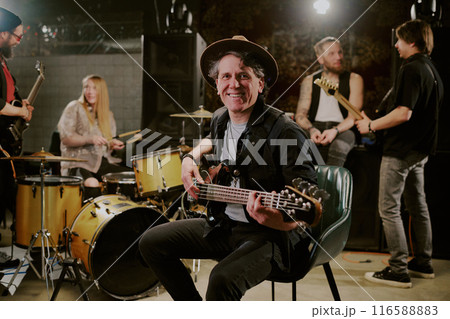 Selective focus portrait of joyful mature Caucasian bassist sitting on chair holding guitar smiling at camera during rehearsal with his band 116588883