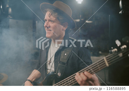 Medium closeup shot of cheerful male rock musician wearing hat playing electric bass guitar on stage Medium closeup shot of cheerful male rock musician wearing hat playing electric bass guitar on stage 116588895