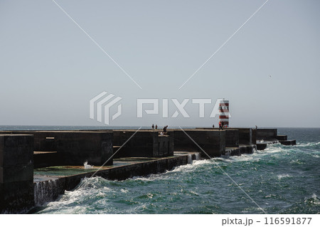 A pier stands in the ocean with a lighthouse in the background A pier stands in the ocean with a lighthouse in the background 116591877