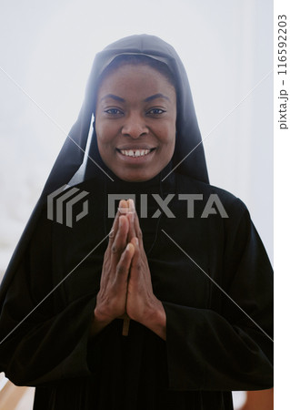 Vertical waist up portrait of happy African American Catholic nun posing for camera with praying hands 116592203