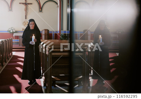 Wide shot of senior Caucasian Catholic nun holding candle in hands standing at nave and her dull reflection on door surface Wide shot of senior Caucasian Catholic nun holding candle in hands standing at nave and her dull reflection on door surface 116592295