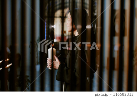 Through gate side view shot of mature Caucasian nun holding candle in hands praying to God 116592313