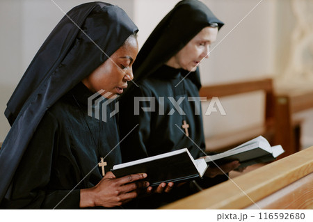 Two ethnically diverse Catholic nuns sitting on pew holding books and saying prayers, medium closeup shot Two ethnically diverse Catholic nuns sitting on pew holding books and saying prayers, medium closeup shot 116592680