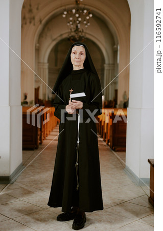 Vertical long shot of mature Caucasian nun holding Bible book in hands standing at narthex in Catholic church, looking at camera Vertical long shot of mature Caucasian nun holding Bible book in hands standing at narthex in Catholic church, looking at camera 116592741