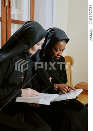 Vertical medium shot of two biracial nuns sitting on wooden bench in Catholic church and discussing Bible texts 116592783
