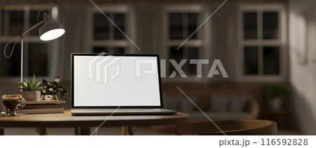 A laptop computer mockup, illuminated by light from a table lamp on a wooden table. 116592828
