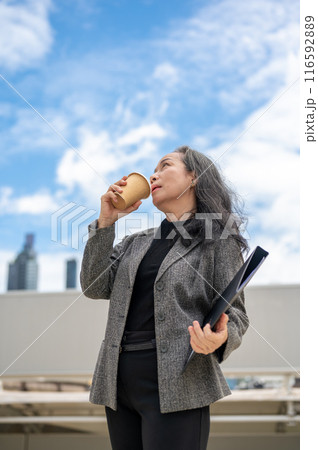 A confident Asian senior businesswoman stands in the urban city surrounded by skyscraper buildings. 116592889