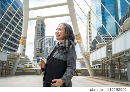 A successful mature Asian businesswoman in a suit is looking away at the sky, standing in the city. 116592902