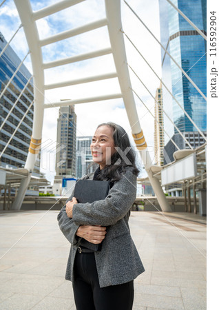 A successful mature Asian businesswoman in a suit is looking away at the sky, standing in the city. 116592904