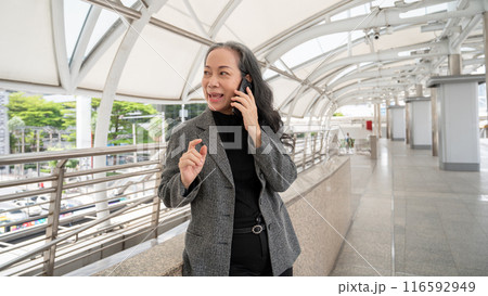 A confident mature Asian businesswoman is talking on the phone while standing on a skywalk. A confident mature Asian businesswoman is talking on the phone while standing on a skywalk. 116592949