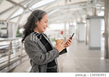 A happy mature Asian businesswoman is reading texts on her smartphone while standing on a skywalk. 116592951