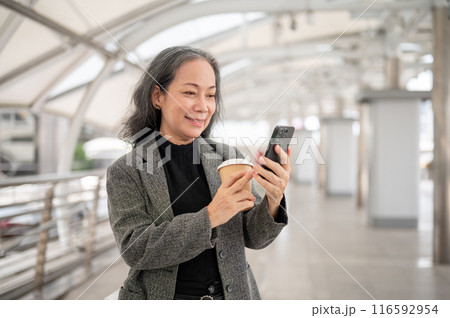 A happy mature Asian businesswoman is reading texts on her smartphone while standing on a skywalk. A happy mature Asian businesswoman is reading texts on her smartphone while standing on a skywalk. 116592954