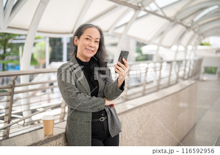 An attractive mature Asian businesswoman stands on a skywalk with a smartphone in her hand. 116592956