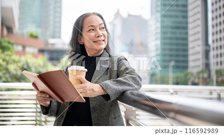 A happy mature Asian businesswoman stands on a skywalk, looking away at the skyscraper buildings. A happy mature Asian businesswoman stands on a skywalk, looking away at the skyscraper buildings. 116592989