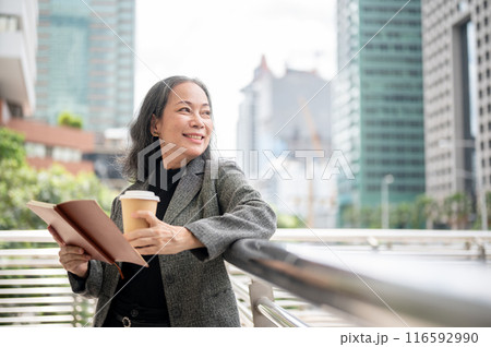 A happy mature Asian businesswoman stands on a skywalk, looking away at the skyscraper buildings. A happy mature Asian businesswoman stands on a skywalk, looking away at the skyscraper buildings. 116592990
