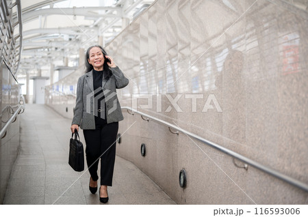 A mature Asian businesswoman is talking on the phone while walking on a skytrain station in the city A mature Asian businesswoman is talking on the phone while walking on a skytrain station in the city 116593006