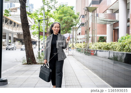 A confident, positive mature Asian businesswoman is walking on a sidewalk in the city. 116593035