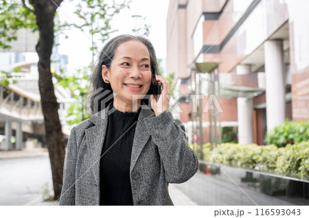 A mature Asian businesswoman is talking on the phone while commuting work, walking on a sidewalk. 116593043
