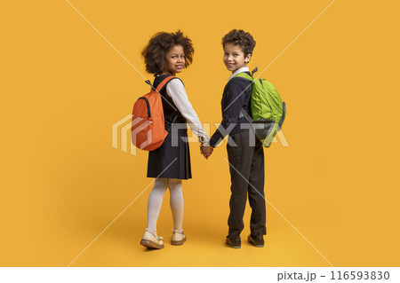 Two African American children, wearing backpacks, are holding hands against a bright yellow background. They appear to be ready for a journey or adventure, back view 116593830