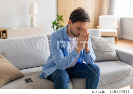 A young man is seated on a grey sofa, looking distressed with his hands covering his face. A smartphone lies nearby, suggesting he may have received upsetting news 116593851