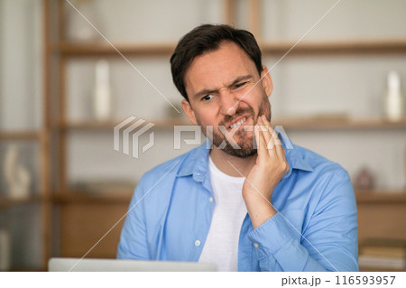 A man sitting in front of a laptop, holding his jaw in pain, indicating he has a toothache. He appears uncomfortable and focused on his dental discomfort. A man sitting in front of a laptop, holding his jaw in pain, indicating he has a toothache. He appears uncomfortable and focused on his dental discomfort. 116593957