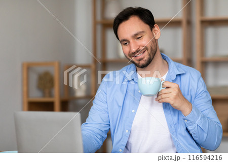 A man is seated at a desk, using a laptop while sipping on a cup of coffee. He appears focused on his work, surrounded by a simple office setting. 116594216