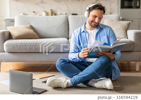 A man is seated on the floor, engrossed in reading a book while simultaneously listening to music through headphones. He appears focused and relaxed in his dual activities. A man is seated on the floor, engrossed in reading a book while simultaneously listening to music through headphones. He appears focused and relaxed in his dual activities. 116594219