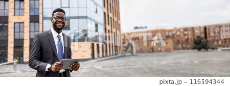Business news. Happy black businessman using digital tablet walking outdoors near modern office building in urban area cityscape, smiling to camera, copy space, web-banner 116594344