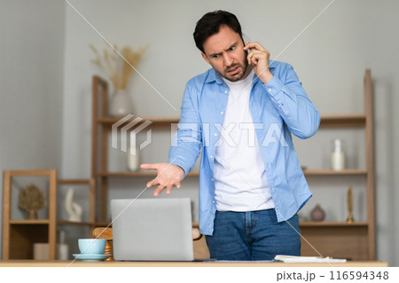 A man stands in front of his laptop in a home office, expressing frustration and confusion during a phone call. His body language, with one hand on his hip and a furrowed brow 116594348