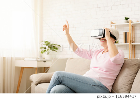 Elderly woman is seated on a couch, wearing a virtual reality headset, engaged in the virtual world, with a hand reaching out in front of her. The room is dimly lit, with the headset glowing blue. 116594417