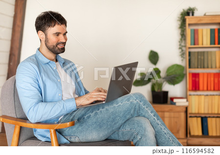 A man is seated in a chair, focused on a laptop computer screen. His hands are typing on the keyboard as he works. The room appears to be well-lit, copy space 116594582