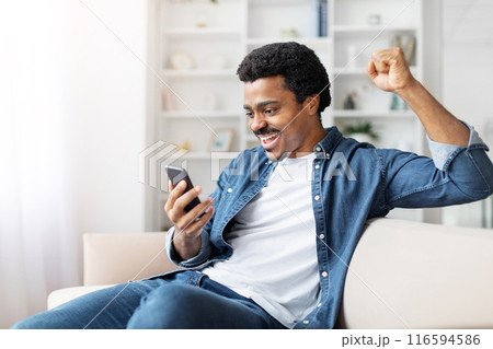 African American man is seated on a couch indoors, holding a cell phone in his hand. He appears focused on the device, with a neutral expression on his face, gesturing 116594586
