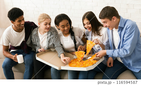 A diverse group of teenagers is sitting together, smiling and enjoying a large pizza. They are casually dressed, indicating a relaxed and social atmosphere 116594587