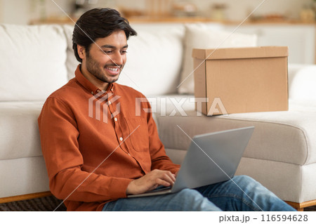 Arab man is sitting on a couch while using a laptop computer. He is focused on the screen, typing and scrolling. The room is cozy with a coffee table and a lamp in the background. Arab man is sitting on a couch while using a laptop computer. He is focused on the screen, typing and scrolling. The room is cozy with a coffee table and a lamp in the background. 116594628
