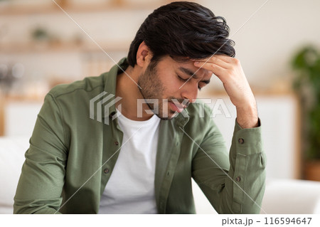 Arab man is seated on a couch, leaning back with his hand on his head. He appears deep in thought or contemplation, with a pensive expression on his face 116594647