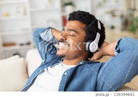African American man is seated on a couch, wearing headphones. He appears focused on listening to music or audio content through the headphones as he relaxes on the couch. 116594681