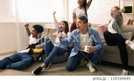 A group of friends is gathered in a cozy living room, expressing joy and excitement. Some are seated on a sofa while others sit on the floor, watching a sports game or a competitive event on TV. 116594695