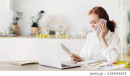 A woman sits at a desk with a laptop open, reviewing documents while talking on her phone. A woman sits at a desk with a laptop open, reviewing documents while talking on her phone. 116594835
