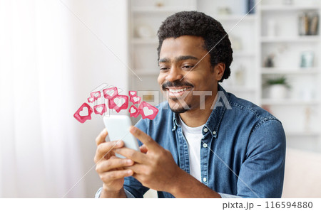 Black man with a joyful expression smiles as he looks at his smartphone. He is clearly pleased with the notifications he is receiving, which are represented by heart-shaped icons floating above Black man with a joyful expression smiles as he looks at his smartphone. He is clearly pleased with the notifications he is receiving, which are represented by heart-shaped icons floating above 116594880
