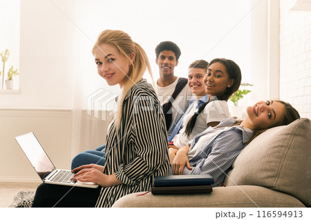 A diverse group of friends sitting closely together on a couch, all focused on a laptop screen. They appear engaged in the content displayed, doing homework A diverse group of friends sitting closely together on a couch, all focused on a laptop screen. They appear engaged in the content displayed, doing homework 116594913