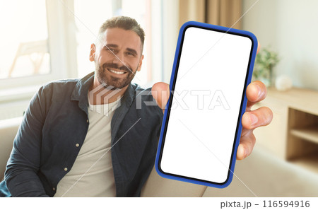 A man is seated on a couch while actively holding up a cell phone in his hand. He appears focused on the devices screen, engrossed in whatever content he is viewing. 116594916