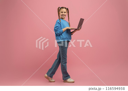 A young girl is walking while holding a laptop in her hands. She appears focused and determined as she moves forward with the device. The background shows a sidewalk with trees lining the path. 116594938