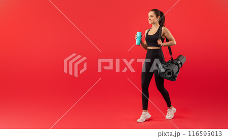 A fit woman dressed in black sportswear confidently strides forward while holding a yoga mat and gym bag. Her white sneakers contrast with the bright red backdrop 116595013