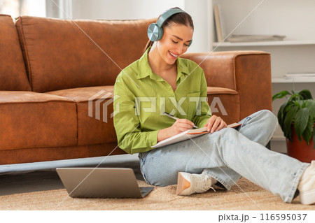 A woman is seated on the floor, using a laptop with headphones on. She appears focused on her screen, possibly working or studying. 116595037