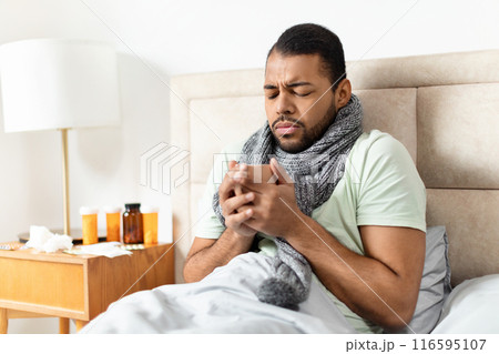 African American young man is sitting up in bed, wearing a light green T-shirt and a thick scarf, looking concerned as he holds a mug with hot drink African American young man is sitting up in bed, wearing a light green T-shirt and a thick scarf, looking concerned as he holds a mug with hot drink 116595107