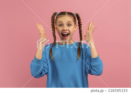 A young girl wearing a bright blue sweater stands against a vibrant pink backdrop, her hands raised in a gesture of surprise and elation A young girl wearing a bright blue sweater stands against a vibrant pink backdrop, her hands raised in a gesture of surprise and elation 116595155