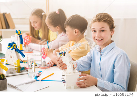 A young boy smiles at the camera while holding a robot during a STEM class. He is sitting at a table with other children who are also working on STEM projects. The classroom is bright and airy 116595174