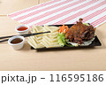 A traditional Asian dish, crispy duck is served with rice sheets, vegetables and dipping sauces on a wooden table, with a stripe towel in the background 116595186